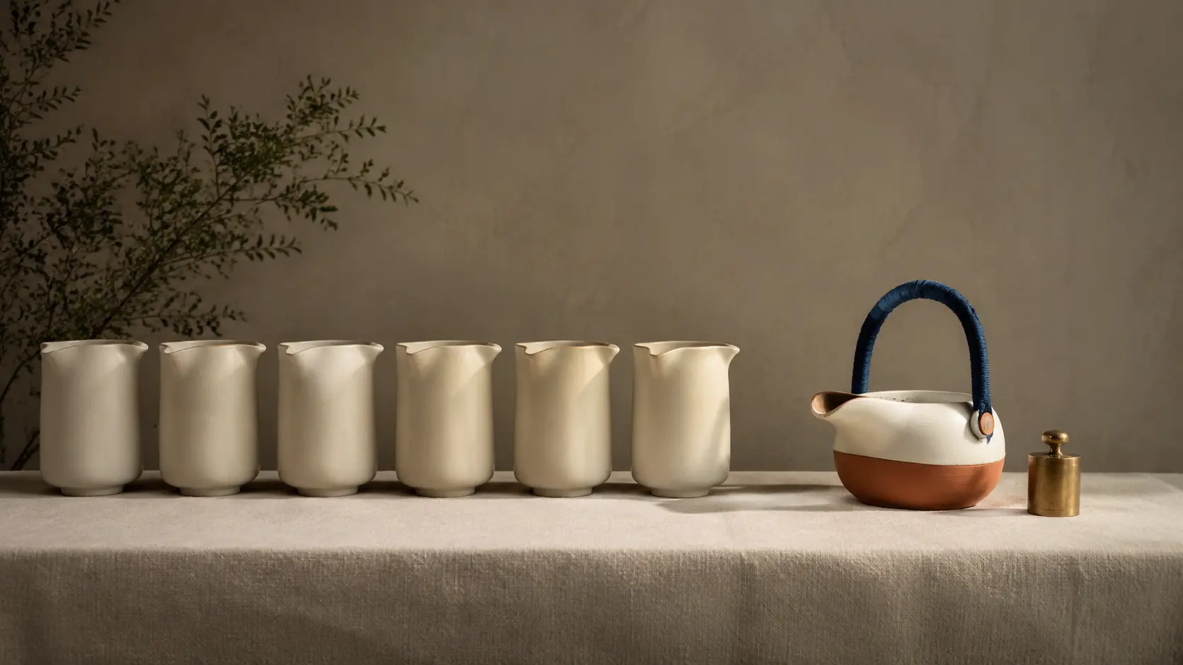 A row of identical cream pitchers on a linen table beside a single cream and terracotta teapot with a blue handle and a small brass weight