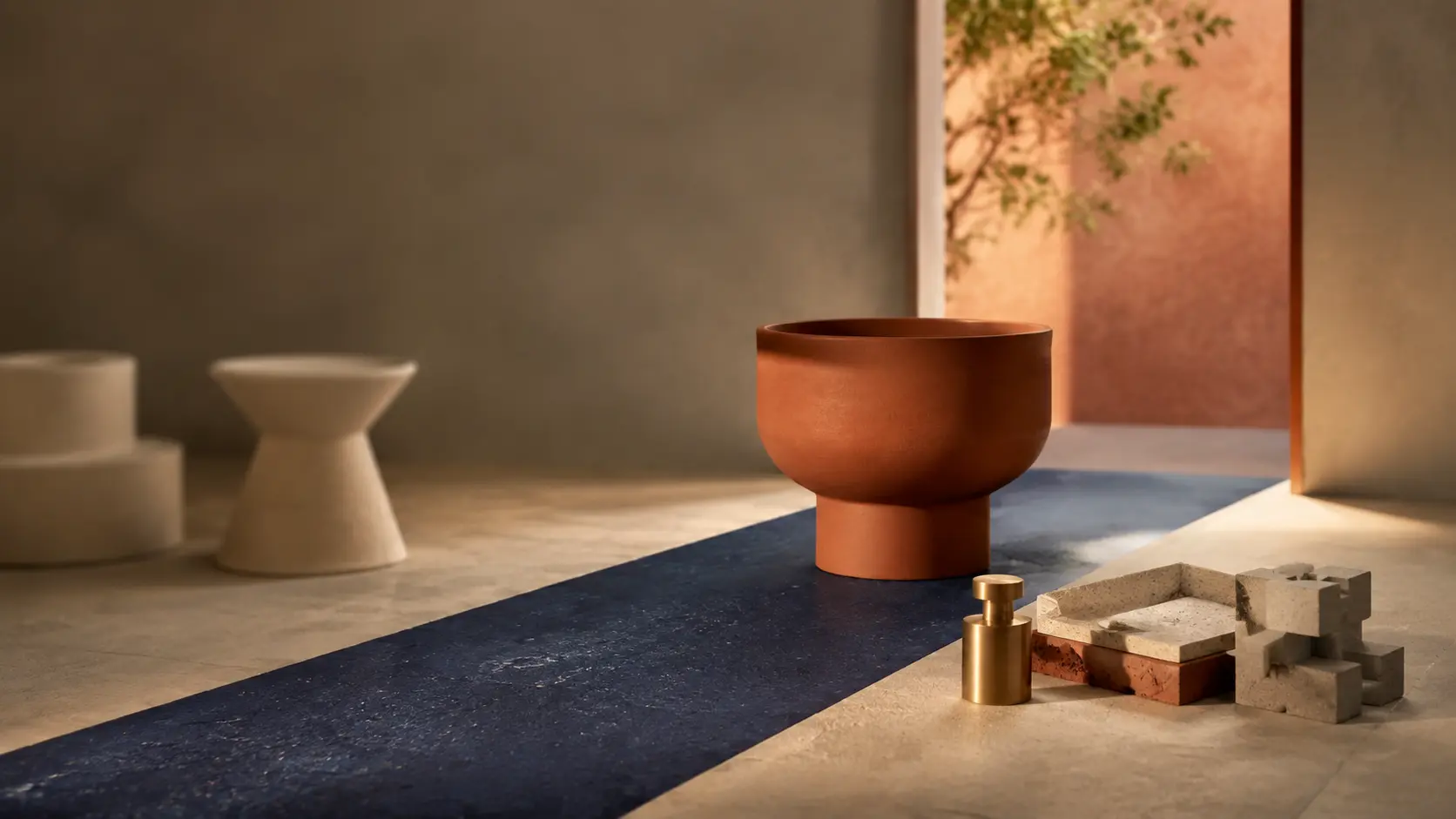 A terracotta bowl on a deep blue runner in a warm plaster room, with a brass weight, stone blocks and a branch of leaves by the doorway