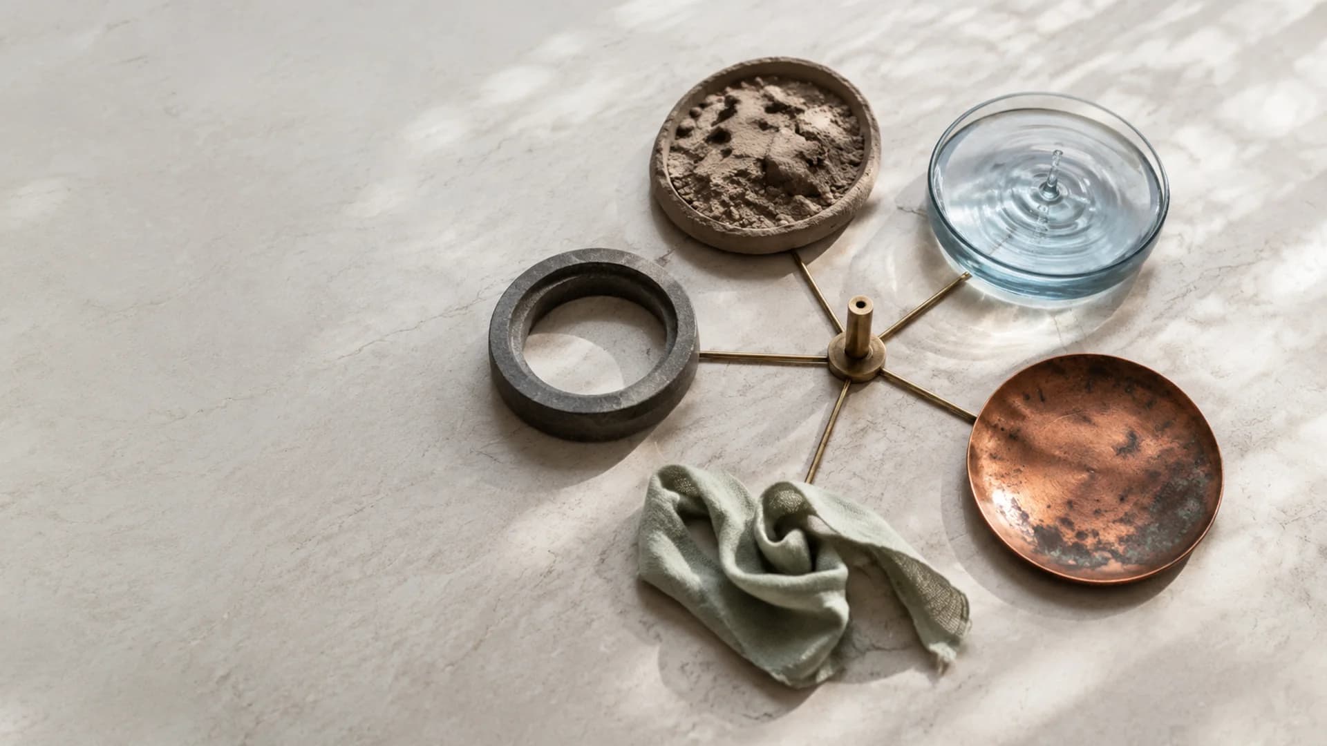 A bowl of earth, a glass dish of water, a copper plate, a stone ring and a folded linen cloth arranged around a small brass post on pale marble